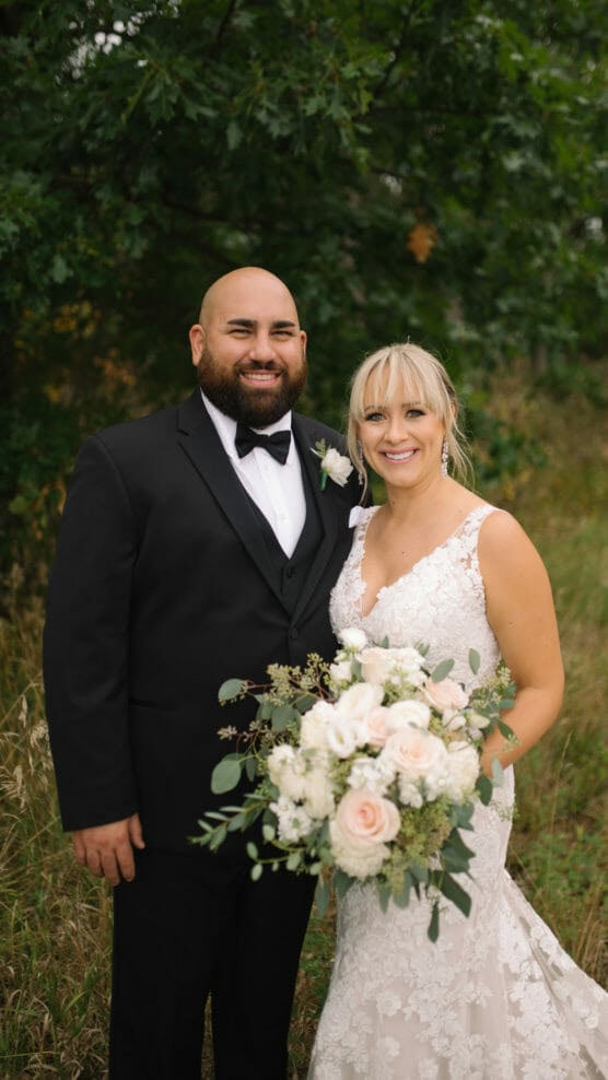 A bride in a white lace dress holding a bouquet of white and blush flowers stands beside a groom in a black tuxedo. They are smiling, posing outdoors in front of green trees and grass. by Justin Salem Meyer