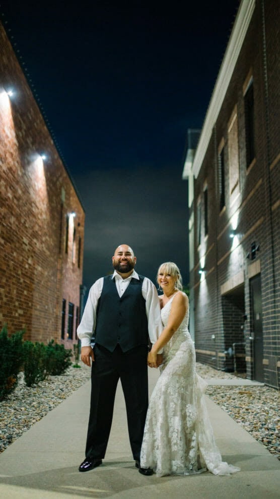 A bride in a lace gown and a groom in a black vest and bow tie stand holding hands and smiling, posing outside at night between two brick buildings. by Justin Salem Meyer