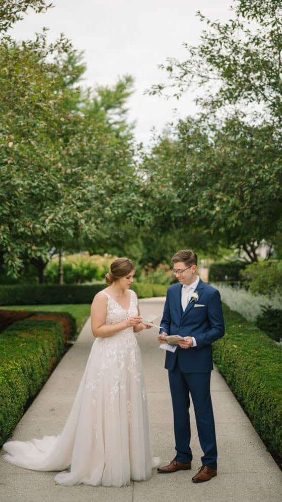 A bride in a white dress and a groom in a blue suit stand on a garden pathway, reading letters to each other, surrounded by greenery and trees. by Justin Salem Meyer