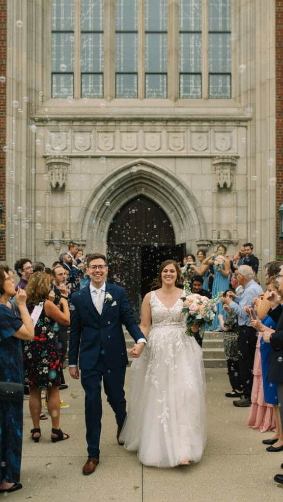 A bride and groom walk hand in hand outside a large stone building, smiling as guests blow bubbles around them. The bride holds a bouquet and wears a white dress, while the groom is in a blue suit. by Justin Salem Meyer