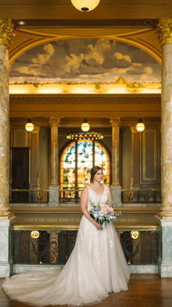 A bride in a white gown holds a bouquet of flowers, standing between marble columns in an elegant hall with ornate decor, a stained glass window, and a painted ceiling in the background. by Justin Salem Meyer
