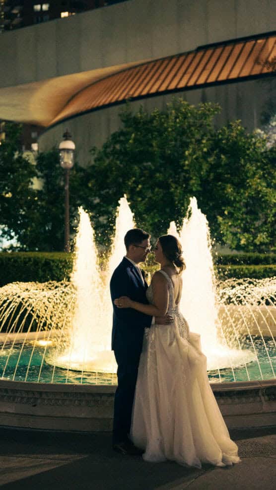 A couple in formal wedding attire embrace and gaze at each other in front of a brightly lit fountain at night, with greenery and a modern building in the background. by Justin Salem Meyer