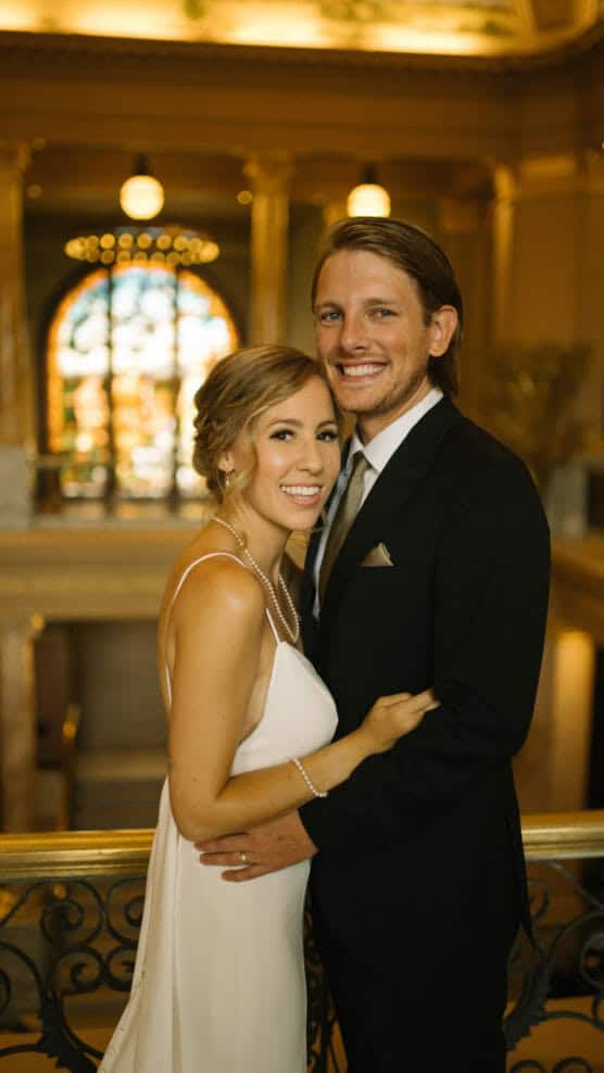 A smiling couple in formal attire pose together indoors; the woman wears a white dress and pearl necklace, and the man wears a black suit and tie. Ornate railings and a grand, warmly lit background are visible. by Justin Salem Meyer