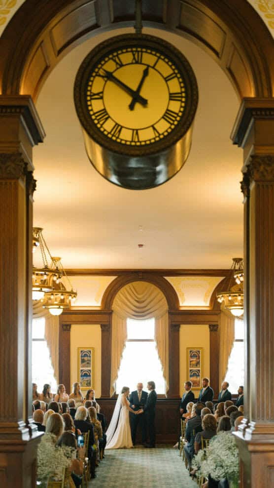 A wedding ceremony takes place in an elegant room with large windows and wooden columns, as guests sit facing a couple and officiant under a large clock displaying 2:15. by Justin Salem Meyer