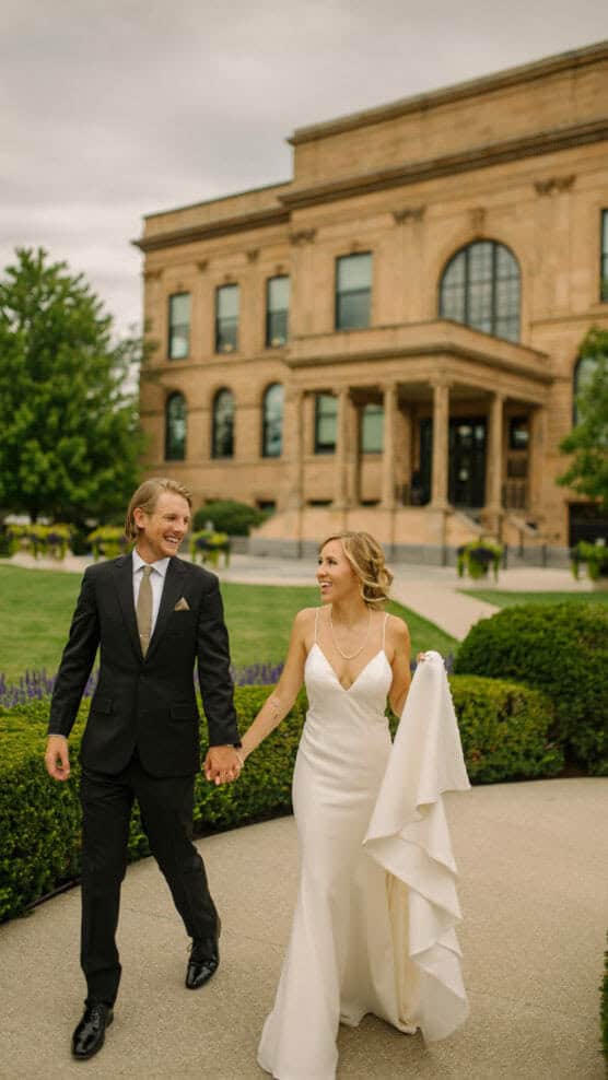 A bride in a white dress and a groom in a black suit walk hand in hand, smiling at each other, on a garden path in front of a large historic building. by Justin Salem Meyer
