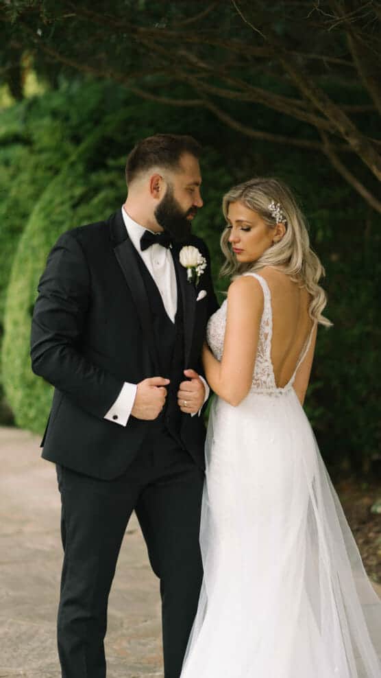 A bride in a white lace dress with an open back stands beside a groom in a black tuxedo. They pose outdoors near greenery, gazing at each other with soft expressions. by Justin Salem Meyer