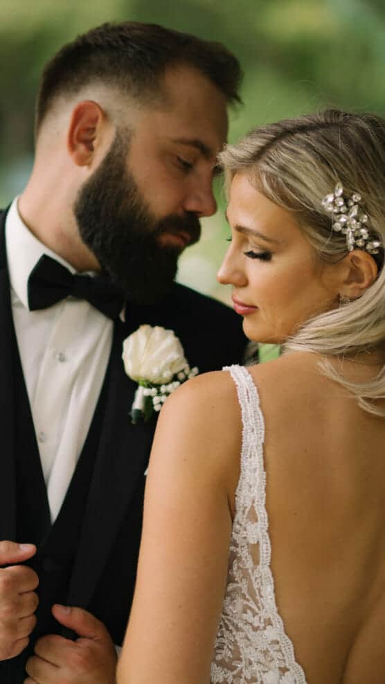 A bride in a lace wedding dress and a groom in a tuxedo with a white rose boutonniere embrace closely, sharing an intimate moment outdoors. by Justin Salem Meyer