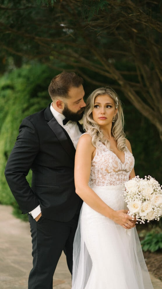 A bride in a white lace gown holds a bouquet of white flowers while standing in front of a groom in a black tuxedo. They pose outdoors with greenery in the background. by Justin Salem Meyer