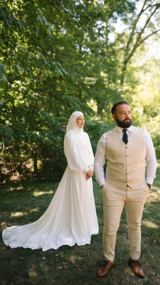 A bride in a white dress and hijab stands calmly behind a groom in a beige suit and black tie. They are outdoors in a sunlit, green, wooded area. The groom faces forward with hands in pockets, while the bride looks at him. by Justin Salem Meyer