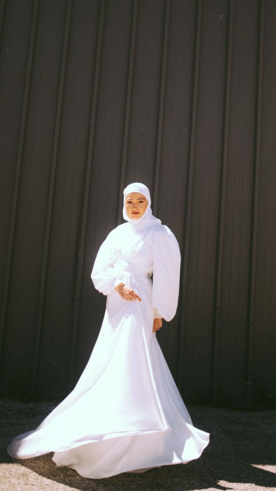 A woman in a flowing white gown and hijab stands in sunlight against a dark vertical-paneled wall, looking calmly at the camera. The dress’s fabric gently flares at the bottom. by Justin Salem Meyer