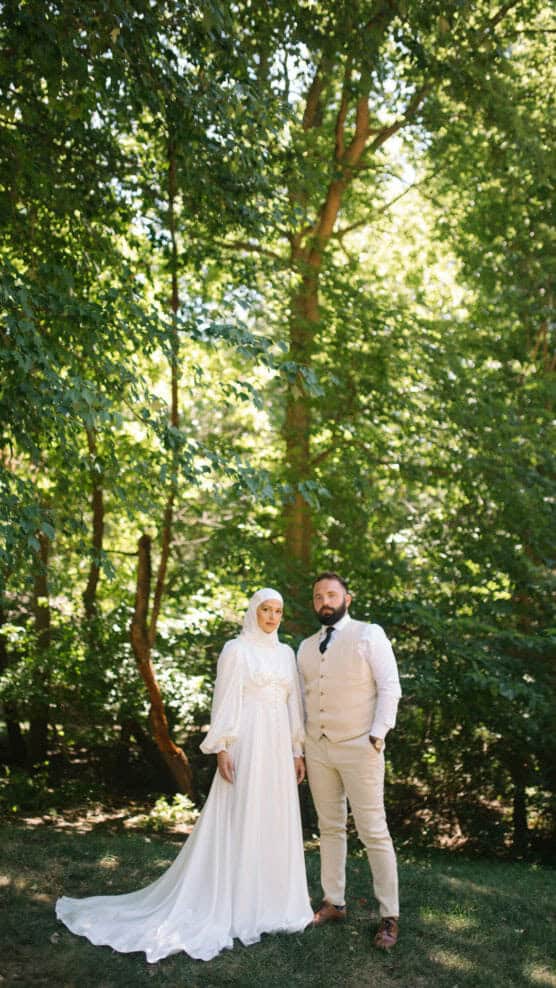 A bride in a white dress and hijab and a groom in a beige suit stand together outdoors in a sunlit, green forest setting. by Justin Salem Meyer