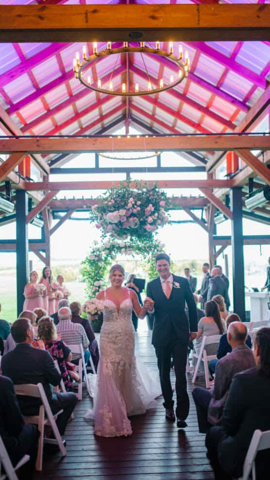 A bride in a white gown and a groom in a dark suit walk down the aisle, smiling and holding hands, as guests seated in a rustic, flower-adorned barn watch and celebrate their wedding ceremony. by Justin Salem Meyer