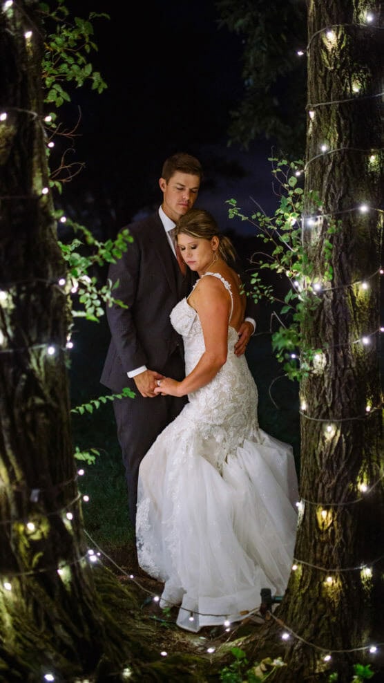 A bride and groom stand closely together between two trees wrapped in string lights at night, holding hands and looking thoughtful, with the bride in a white gown and the groom in a dark suit. by Justin Salem Meyer