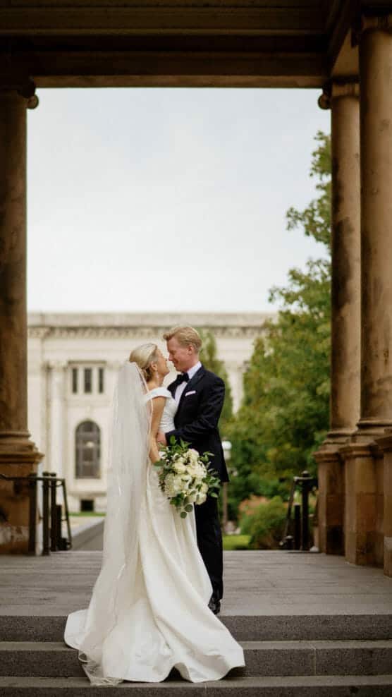 A bride and groom stand close together on stone steps, gazing into each other’s eyes. The bride holds a bouquet of white flowers, and both wear formal wedding attire. Large pillars and a historic building are in the background. by Justin Salem Meyer