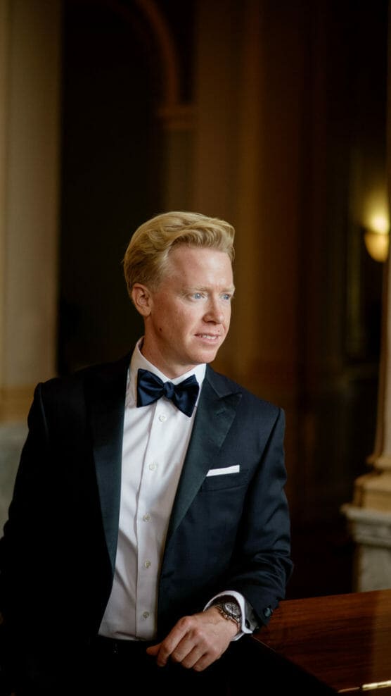 A man with blond hair in a black tuxedo and bow tie stands indoors, leaning on a wooden railing and looking to the side, with soft light illuminating his face. by Justin Salem Meyer