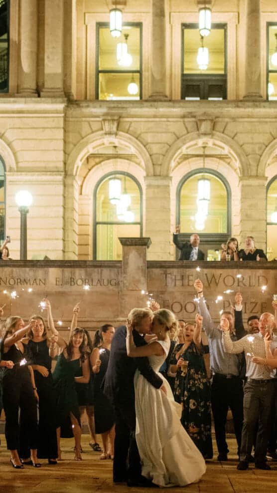 A bride and groom share a kiss outside a grand building at night, surrounded by guests holding sparklers and celebrating. People cheer and wave sparklers, while others watch from a balcony above. by Justin Salem Meyer