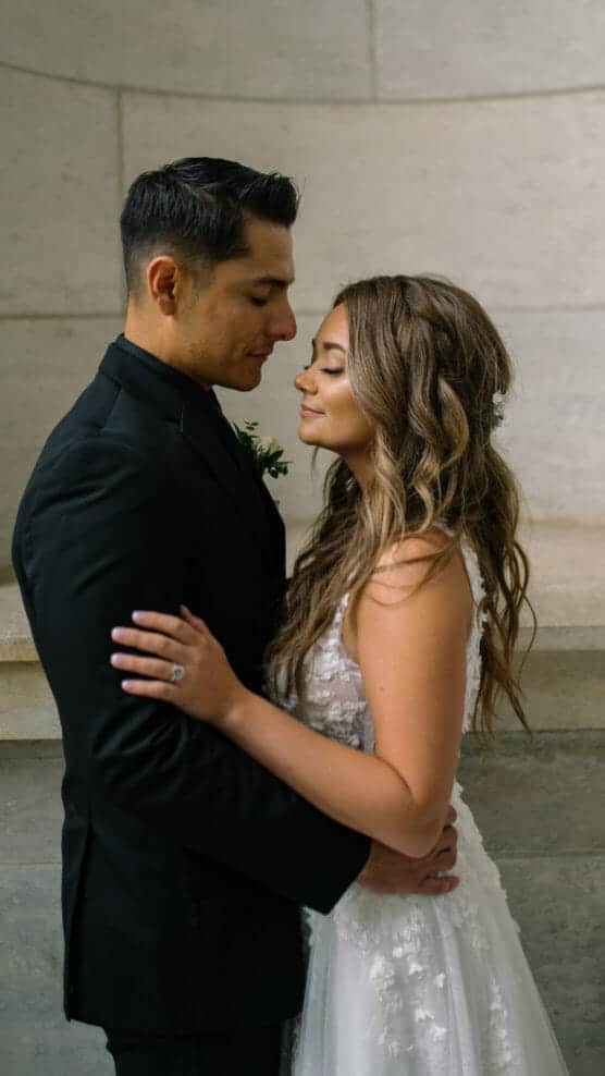 A bride and groom, dressed in a white wedding gown and black suit, stand closely embracing each other and gazing lovingly with closed eyes against a neutral stone background. by Justin Salem Meyer