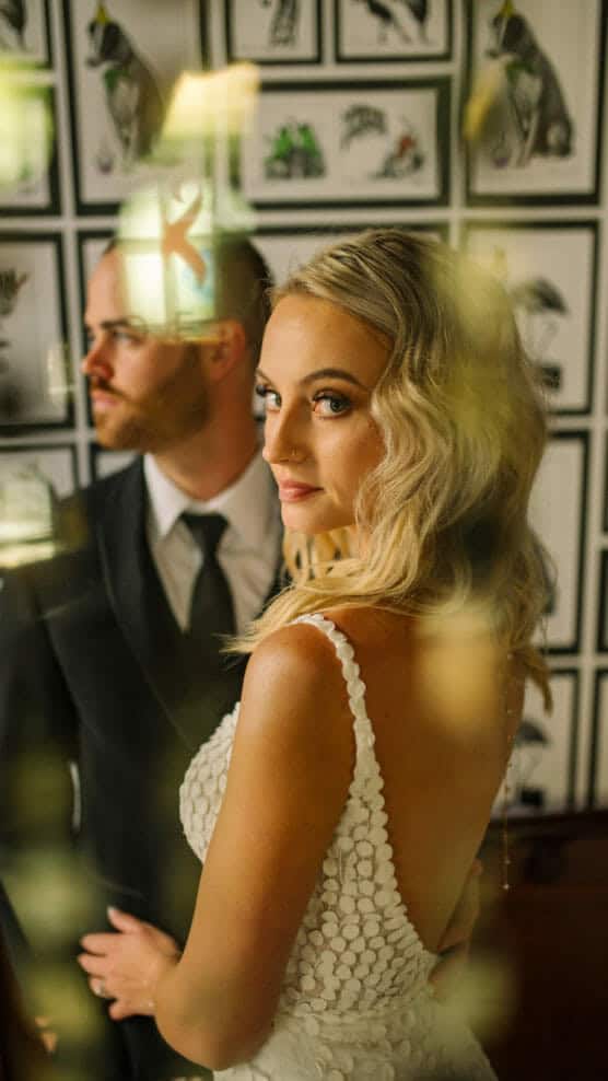 A bride in a white dress with textured details stands beside a groom in a black suit and tie. They pose indoors, with framed artwork and soft, warm light in the background. The bride looks at the camera. by Justin Salem Meyer