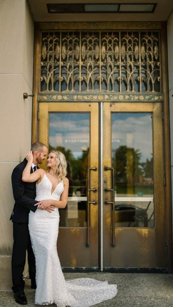 A bride in a white, textured gown and a groom in a black suit embrace and smile in front of ornate, tall brass doors with decorative metalwork. by Justin Salem Meyer
