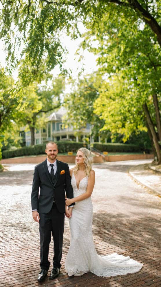 A bride in a white dress and a groom in a dark suit stand hand-in-hand on a sunlit, tree-lined brick road, with greenery and a large house in the background. by Justin Salem Meyer