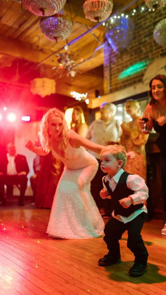 A bride in a white dress and a young boy in a vest and dress shirt dance enthusiastically on a wooden floor at a festive indoor celebration, with colorful lights and people watching in the background. by Justin Salem Meyer