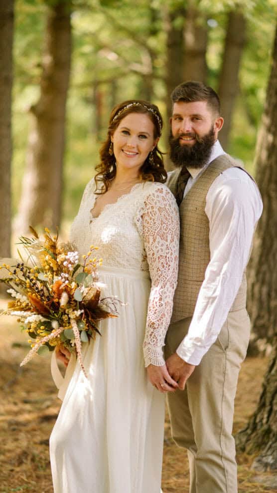 A bride in a long-sleeved lace dress holds a rustic bouquet and stands beside a groom in a vest and tie. They smile, holding hands, surrounded by trees in a sunlit forest setting. by Justin Salem Meyer