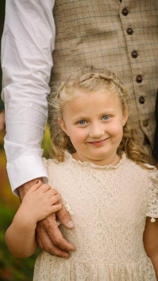 A young girl with blonde hair in a cream lace dress smiles while holding an adults hand. The adult, wearing a white shirt and plaid vest, stands protectively behind her. The background is softly blurred greenery. by Justin Salem Meyer