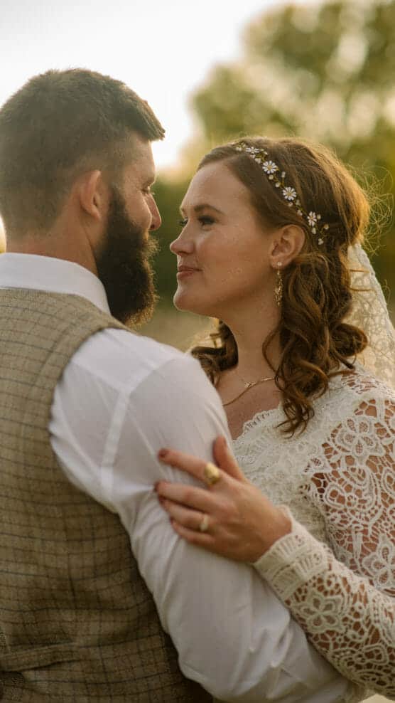 A bride and groom embrace outdoors, looking into each other’s eyes. The bride wears a lace dress and floral headband, while the groom wears a checked vest and white shirt. Sunlight and trees are visible in the background. by Justin Salem Meyer