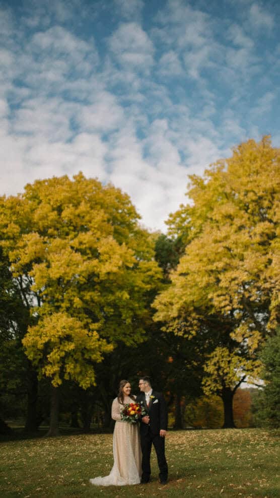 A bride and groom stand together on a grassy field, surrounded by tall trees with autumn leaves. The bride holds a bouquet, and both smile at each other under a blue sky with scattered clouds. by Justin Salem Meyer