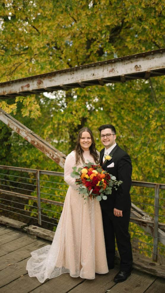 A bride in a light pink gown holds a vibrant bouquet of red and yellow flowers while standing next to the groom in a black suit and yellow tie. They smile on a rustic wooden bridge with green and yellow trees in the background. by Justin Salem Meyer