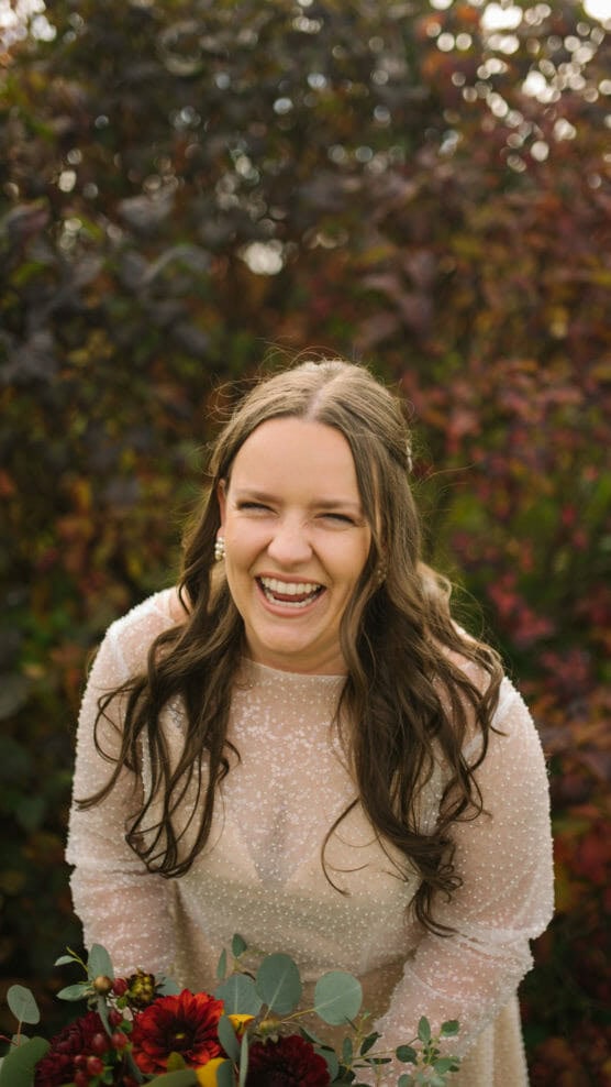 A woman with long brown hair in a white dress smiles and laughs outdoors, holding a bouquet of red flowers and greenery, with colorful autumn foliage in the background. by Justin Salem Meyer