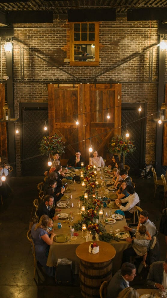 A group of people sit around a long, decorated dinner table in a rustic, warmly lit venue with brick walls, wooden accents, and hanging lights above. The atmosphere is festive and cozy. by Justin Salem Meyer