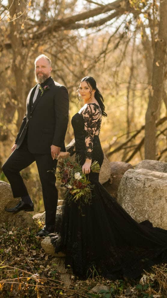 A couple poses outdoors on rocks; the man wears a black suit and the woman wears a long black lace gown, holding a bouquet. Leafless trees and warm sunlight create an autumnal background. by Justin Salem Meyer