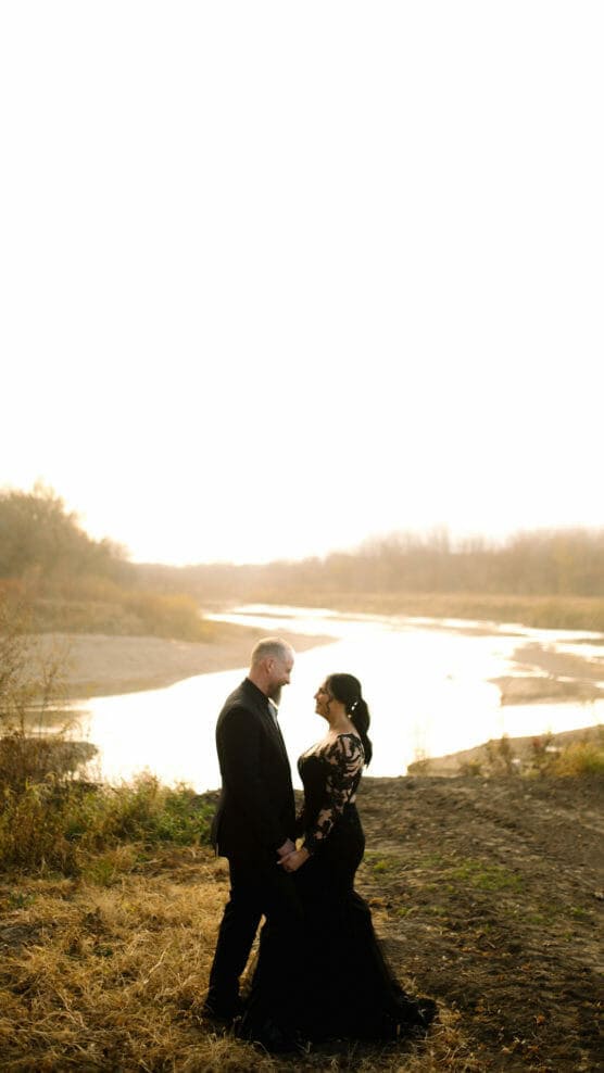 A couple dressed in formal black attire stands hand in hand, gazing at each other by a river at sunset, surrounded by autumn foliage and soft golden light. by Justin Salem Meyer