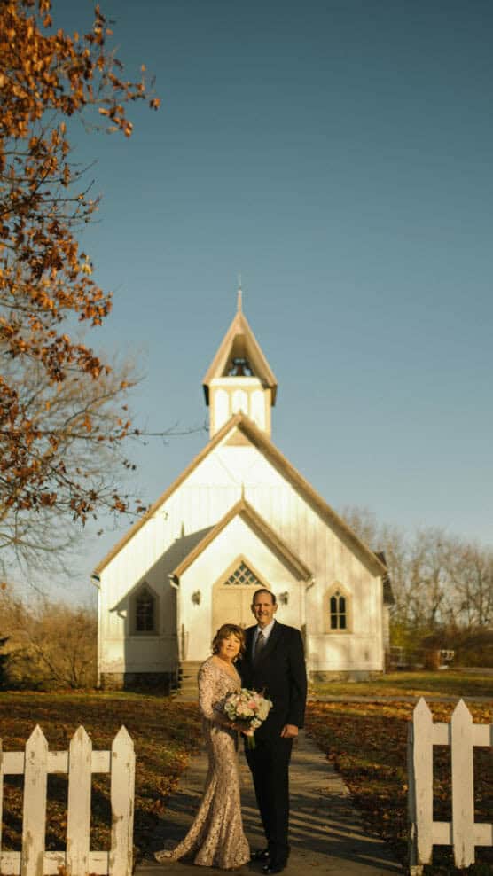 A couple dressed formally stands smiling in front of a white church with a steeple. The sky is clear, and autumn leaves are visible on a tree to the left. A white picket fence frames the foreground. by Justin Salem Meyer