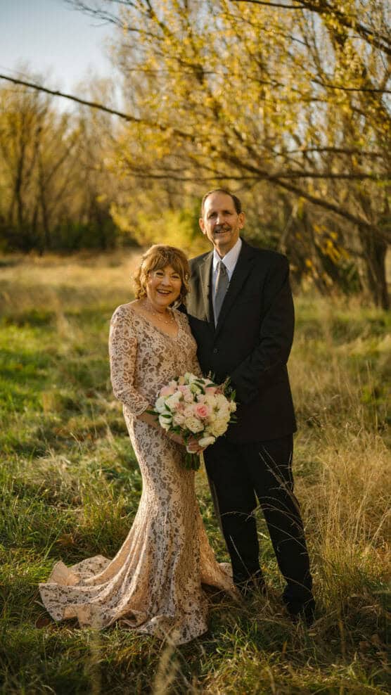 A smiling couple stands outdoors in a grassy field with autumn trees. The woman wears a long lace dress and holds a bouquet of flowers, while the man wears a dark suit and tie. They pose closely together in warm sunlight. by Justin Salem Meyer