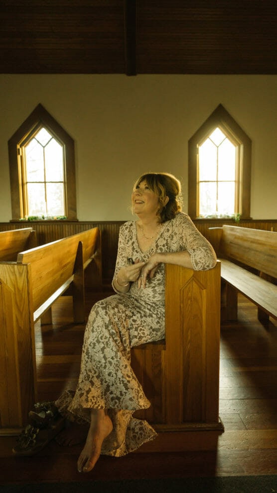 A woman in a long, lace dress sits barefoot and smiling on a wooden pew inside a sunlit church, with two arched windows in the background. Sandals are on the floor beside her. by Justin Salem Meyer
