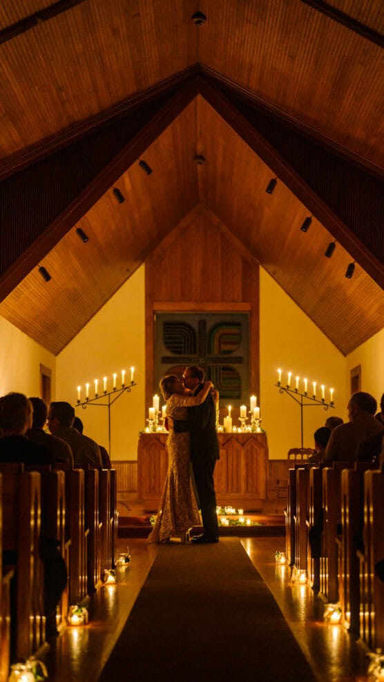 A couple shares a romantic embrace at the altar in a warmly lit chapel, surrounded by candles and guests seated in pews, creating an intimate, cozy wedding atmosphere. by Justin Salem Meyer