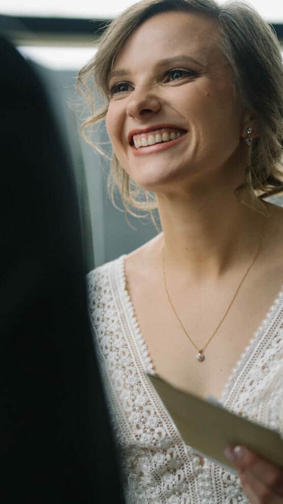 A smiling bride in a white lace dress holds a piece of paper, looking up with joy during a wedding ceremony. by Justin Salem Meyer