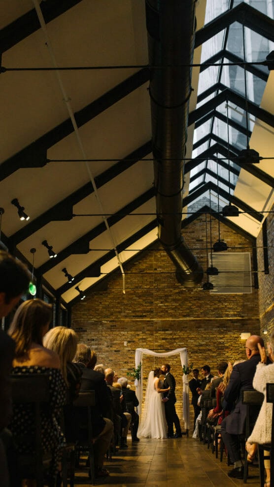 A wedding ceremony takes place indoors, with a couple standing at the altar under a white arch. Guests sit on both sides, facing the couple, in a room with brick walls and a high, glass ceiling. by Justin Salem Meyer