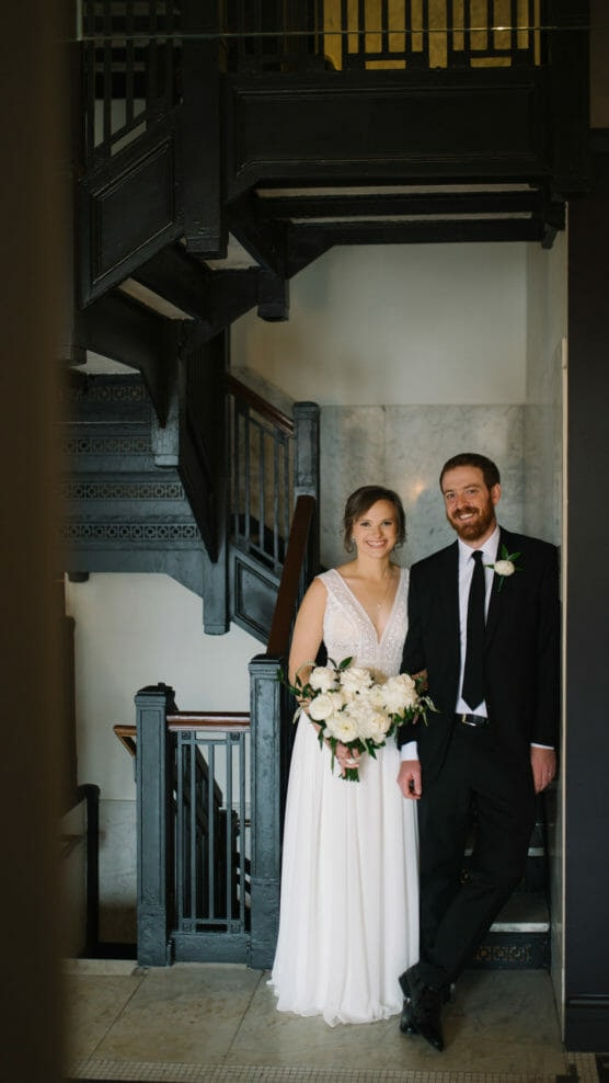 A bride in a white dress holding a bouquet of white flowers stands next to a groom in a black suit and tie. They are smiling and posing under a dark staircase inside a building. by Justin Salem Meyer