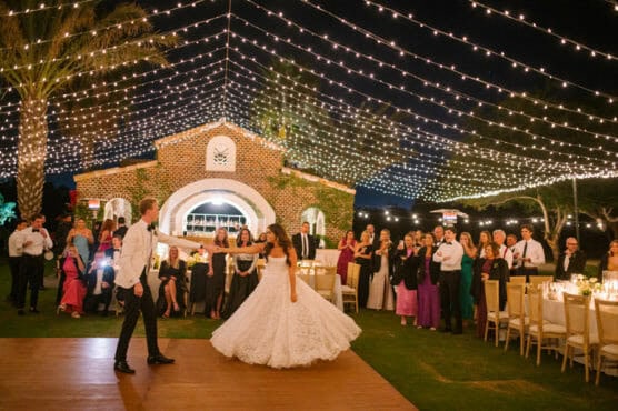 A bride and groom dance under strings of lights at an outdoor wedding reception, surrounded by seated and standing guests near a lit building at night. by Justin Salem Meyer