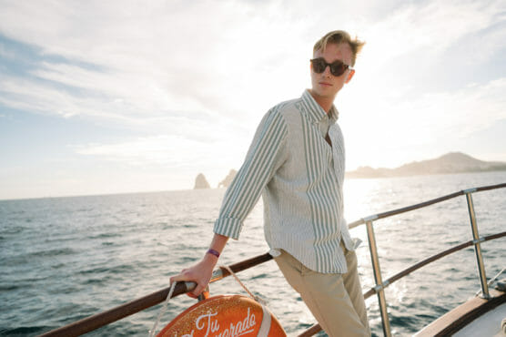 A young man wearing sunglasses and a striped shirt leans on a boat railing, looking away from the camera. The sun is shining brightly, and the ocean with distant islands is visible in the background. by Justin Salem Meyer