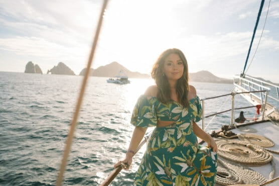 A woman in a green floral dress stands on a boat, holding a railing, with the sun shining behind her and rocky islands visible in the ocean background. by Justin Salem Meyer