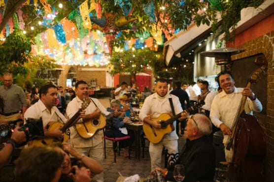 A lively mariachi band performs for diners at a festive, colorful outdoor restaurant decorated with string lights and papel picado banners. Guests enjoy the atmosphere, food, and music under a tree-filled patio. by Justin Salem Meyer