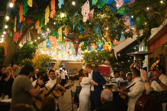 A couple kisses in the center of a lively outdoor celebration decorated with colorful papel picado banners and string lights, as musicians play guitars and guests look on and take photos. by Justin Salem Meyer