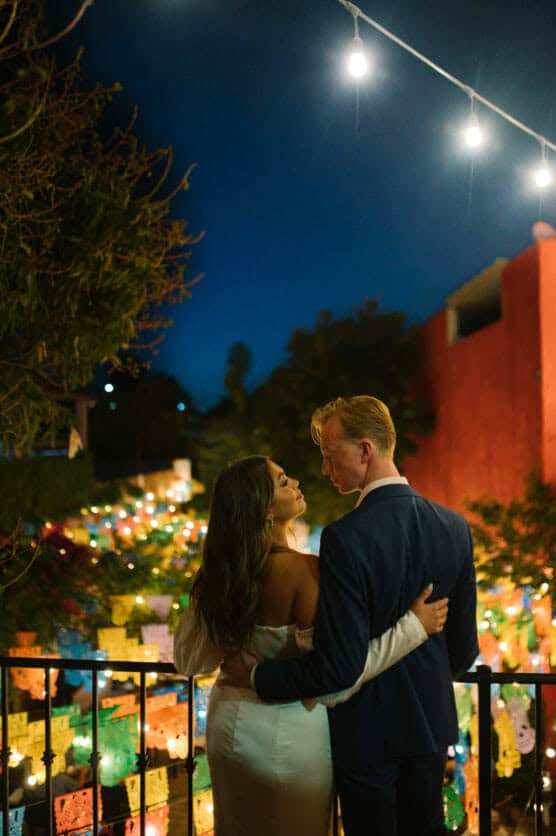 A couple in formal attire embraces on a balcony at night, surrounded by colorful string lights and festive papel picado banners, with warm lighting and a vibrant, romantic atmosphere. by Justin Salem Meyer