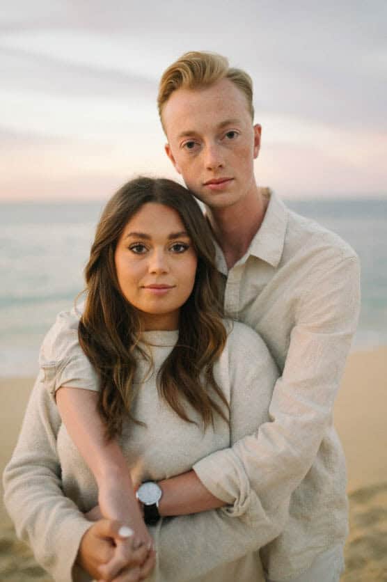 A young couple stands close on a beach at sunset. The man, in a light shirt, gently hugs the woman from behind. Both have serious expressions and look directly at the camera. The ocean and sky are softly blurred in the background. by Justin Salem Meyer