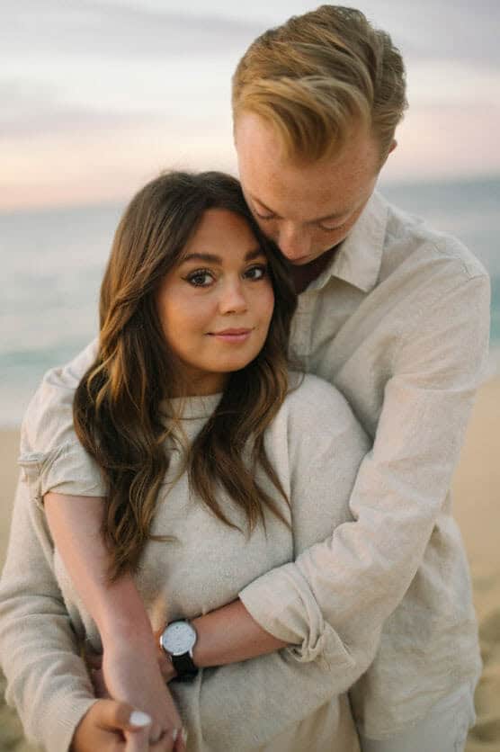 A couple embraces on a beach at sunset. The woman faces the camera with a soft smile, while the man stands behind her, hugging her and looking down. Both wear light-colored, casual clothing. The ocean is blurred in the background. by Justin Salem Meyer