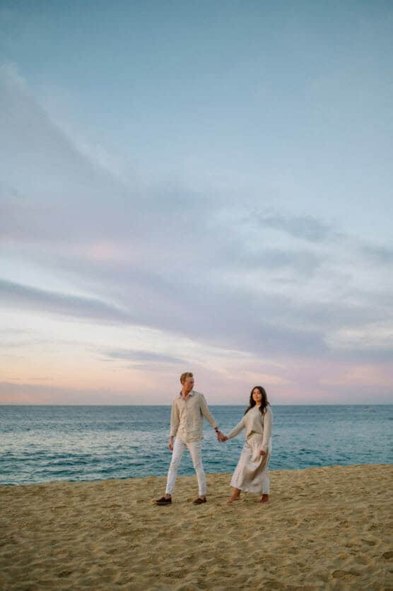 A couple dressed in light-colored clothes walks hand in hand along a sandy beach at sunset, with the ocean and a colorful sky in the background. by Justin Salem Meyer